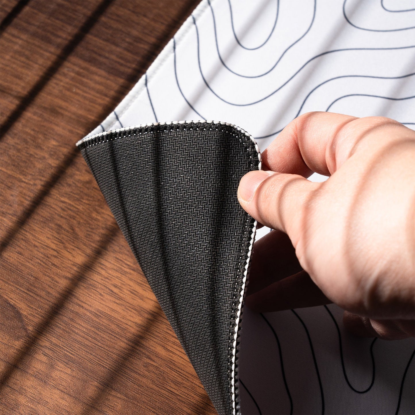 Black desk mat corner lifted by hand; stitched border and wave line pattern visible.