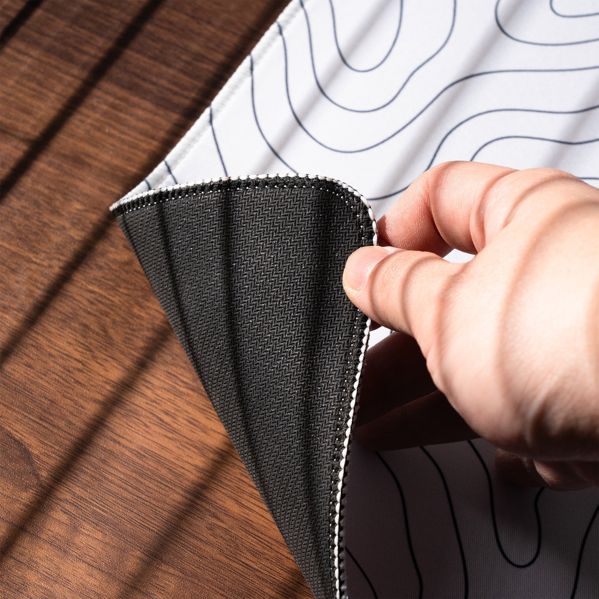 Black desk mat corner lifted by hand; stitched border and wave line pattern visible.