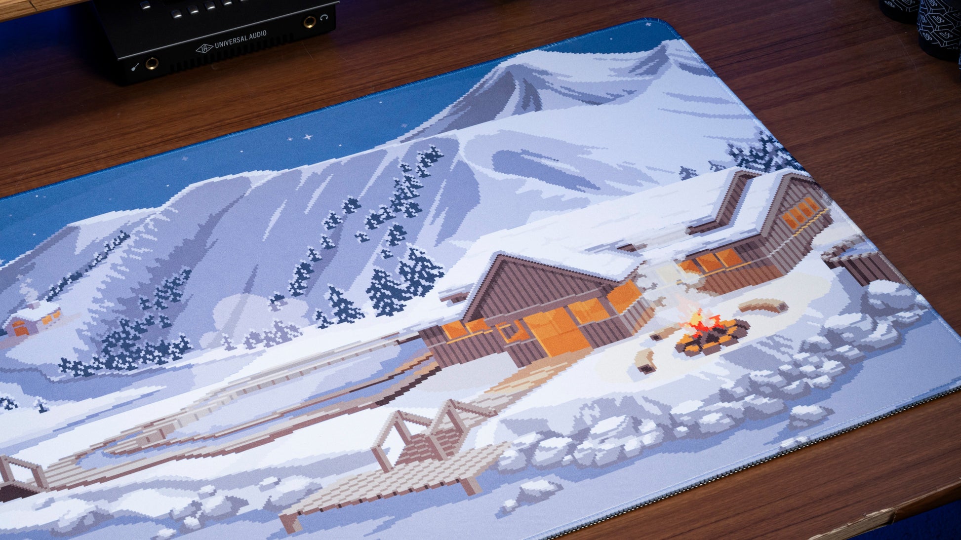 Winter desk mat close-up with snowy mountains, chalet rooftops, and pine trees.